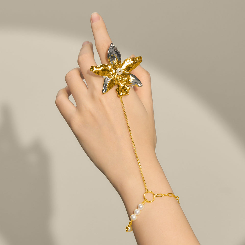 Risis 1974 Odontoglossum Hand Chain - A hand is adorned with a golden flower-shaped ring with silver and gold petals and pearl detail, connected by a chain to a bracelet with more pearls.