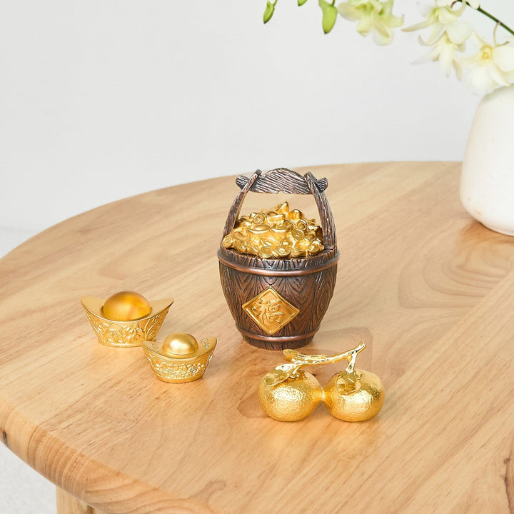 Risis Abundance Money Barrel - A wooden-textured basket filled with gold ingots sits beside golden Chinese ingot-shaped decorations on a polished wood surface, conveying a sense of prosperity and cultural artistry.
