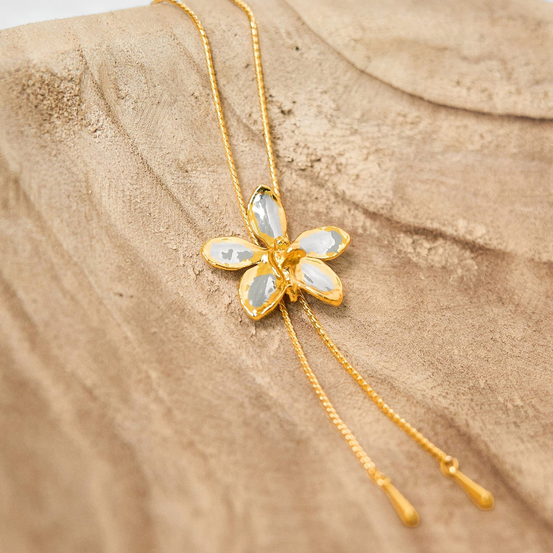 Risis Aeridovanda Vieng Ping Orchid Slider Necklace (PG) - A golden necklace with a central flower-shaped pendant featuring five glossy, translucent petals set in a shiny metal forms the centerpiece, accompanied by two slender dangling chains.