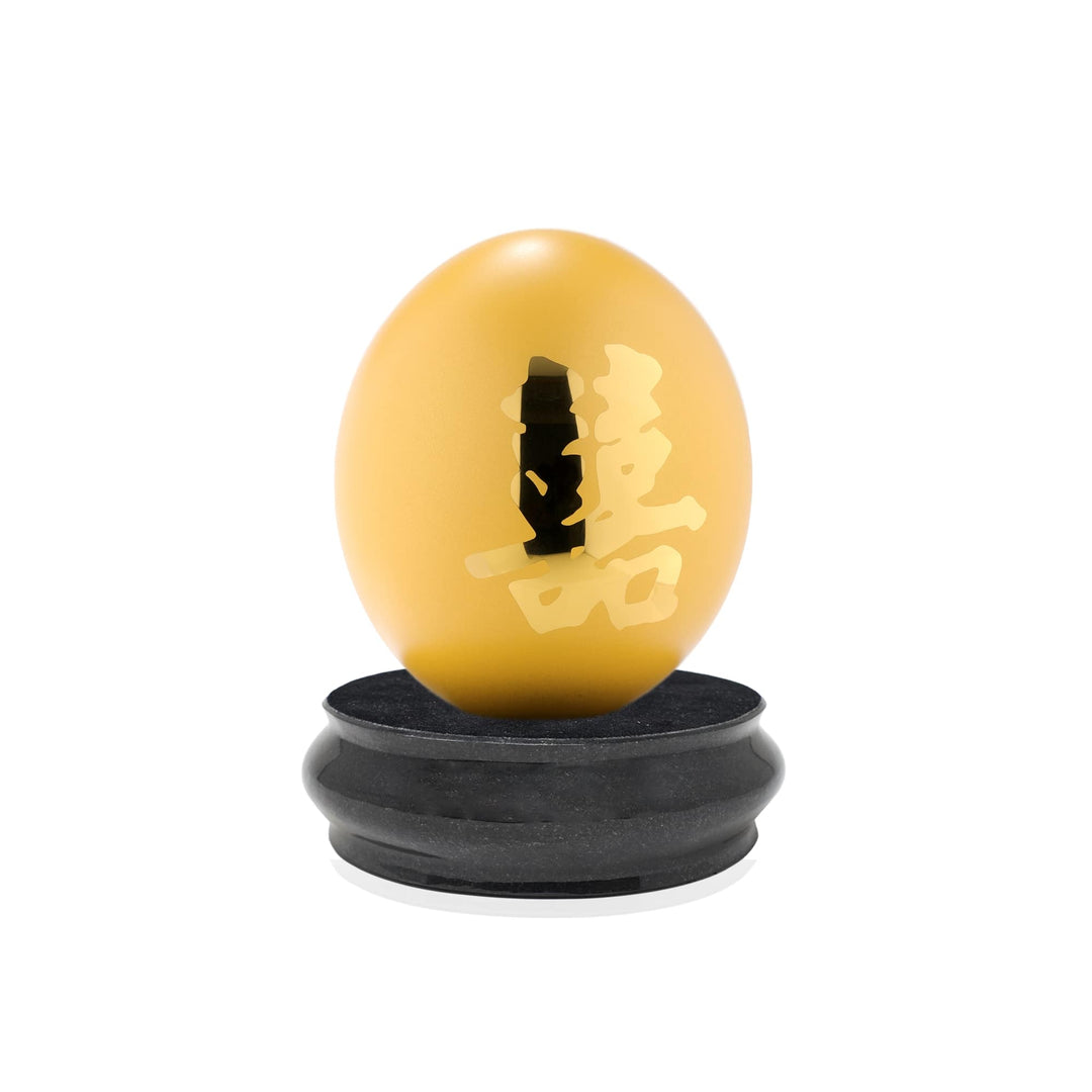 Risis Auspicious Egg II - Xi (Large) - A golden egg-shaped object with intricate carvings sits atop a black circular stand, exhibiting an ornate and polished appearance.