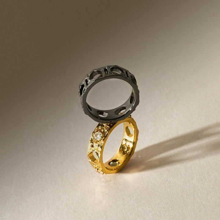 Risis Entwined Rattan Ring - Two rings are displayed, one in gold with textured embellishments and the other in a dark metal with cut-out patterns, both reflecting hues and casting shadows.