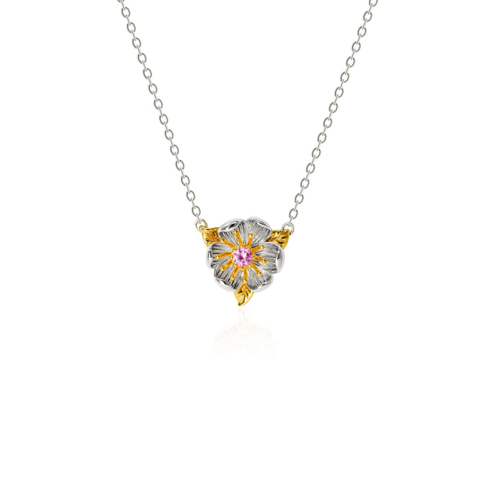 Risis Magnolia Blossom Necklace - A delicate necklace with a silver chain holds a heart-shaped pendant. The pendant features gold and silver petals and a central pink gemstone.