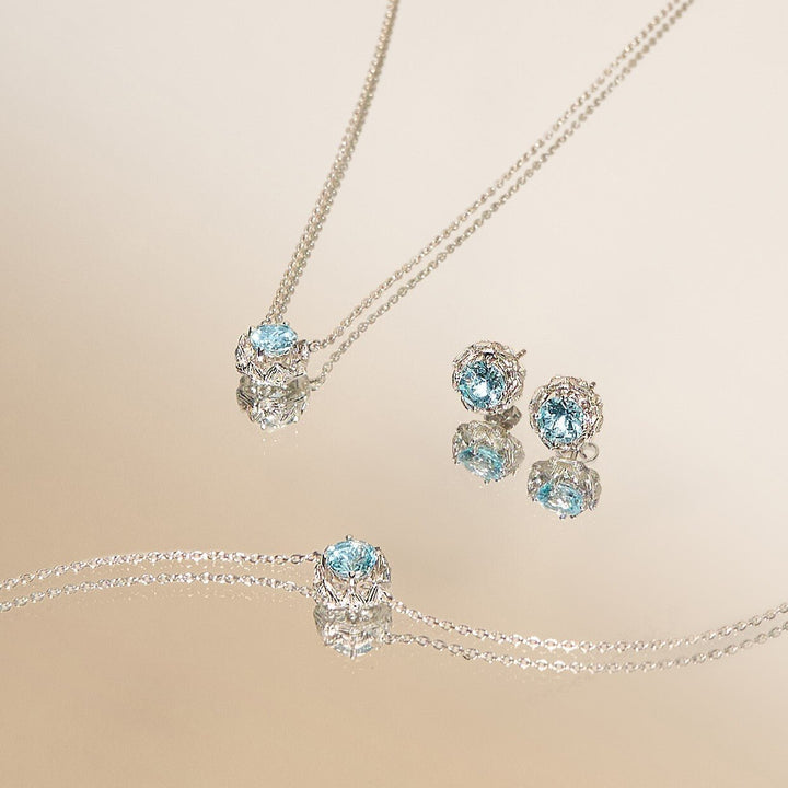 Risis Midori Necklace With Sky Blue Topaz (RH) - A set of silver jewelry with light blue gemstones surrounded by smaller clear crystals, including a pendant necklace and matching stud earrings, displayed on a neutral background.
