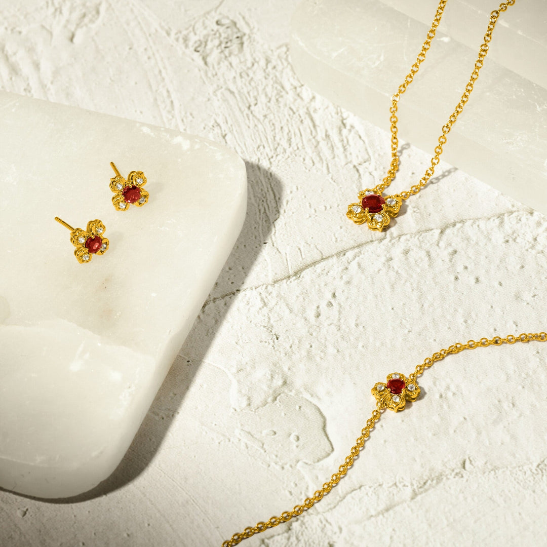 Risis Petite Peranakan Kebaya Necklace With Garnet - Golden jewelry with red gemstones and small crystals rests delicately on a textured surface, displaying a symmetrical floral design.