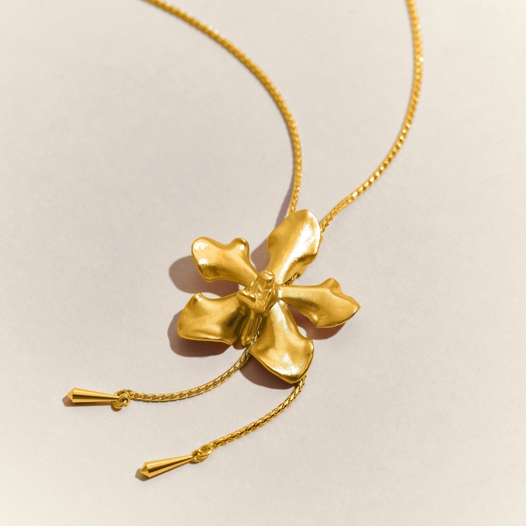 Risis Vanda Limbata Orchid Slider G - A golden necklace with a large, shiny, sculpted flower pendant, detailed with textured petals, and two small golden bars dangling from delicate chains.