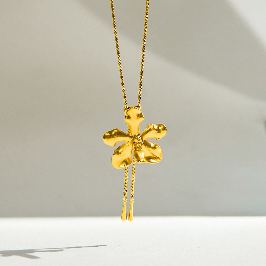 Risis Vanda Limbata Orchid Slider Necklace (G) - A golden necklace with a shiny, textured five-petaled flower pendant, accented by a center gemstone and a delicate hanging detail.