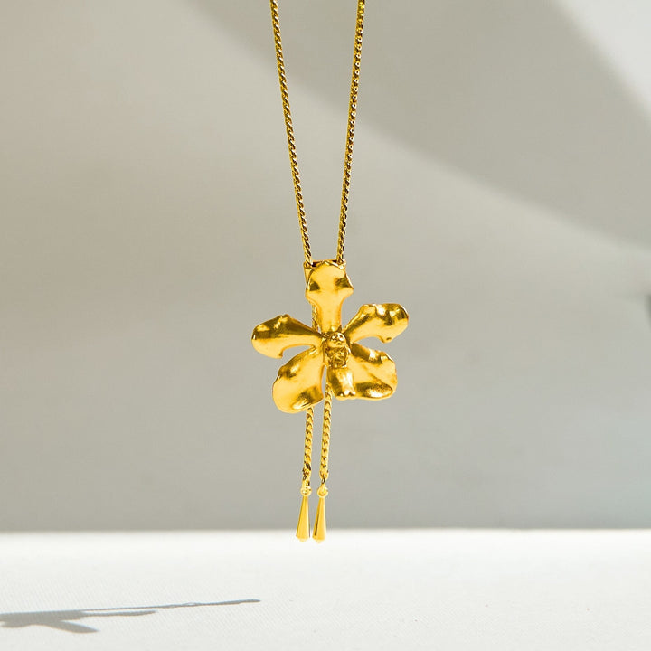 Risis Vanda Limbata Orchid Slider Necklace (G) - A golden necklace with a shiny, textured five-petaled flower pendant, accented by a center gemstone and a delicate hanging detail.