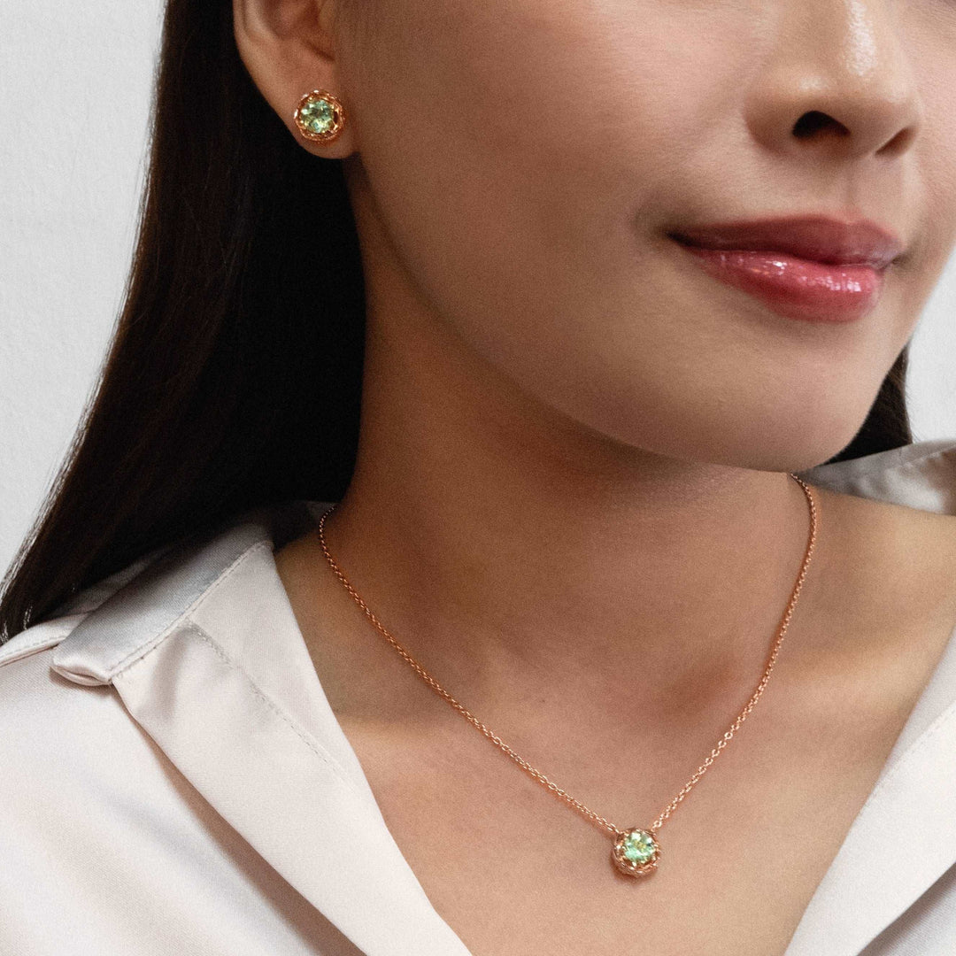 Risis Midori Earrings with Prasiolite (RG) - A woman showcases a rose-gold necklace with a single, sparkling green gem pendant and matching stud earrings, conveying elegance and simplicity.