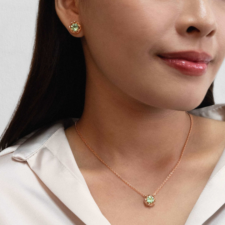 Risis Midori Earrings with Prasiolite (RG) - A woman showcases a rose-gold necklace with a single, sparkling green gem pendant and matching stud earrings, conveying elegance and simplicity.