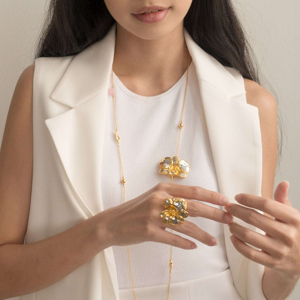 Risis Merry Blossom Necklace - A woman showcases golden jewelry: A delicate necklace with soft pink and clear gems, and rings and brooch with floral motifs and glittering yellow stones.