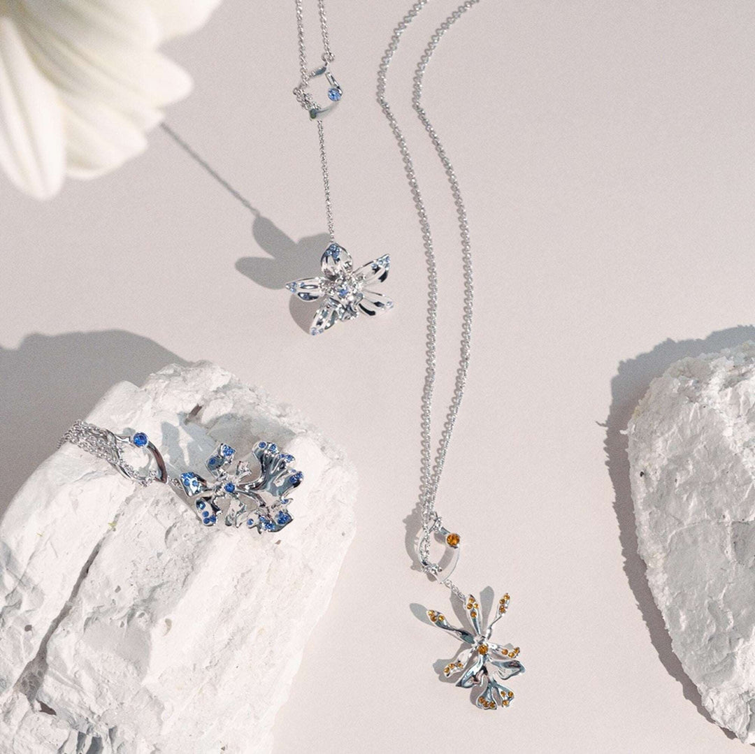 Risis Dazzle With Orchids Necklace - Oncidium Inspired Orchid with Sapphire (Sept) - A collection of silver jewelry with blue and clear gemstones, designed in floral patterns, elegantly drapes over stark white stones, creating a contrast of natural and crafted beauty.