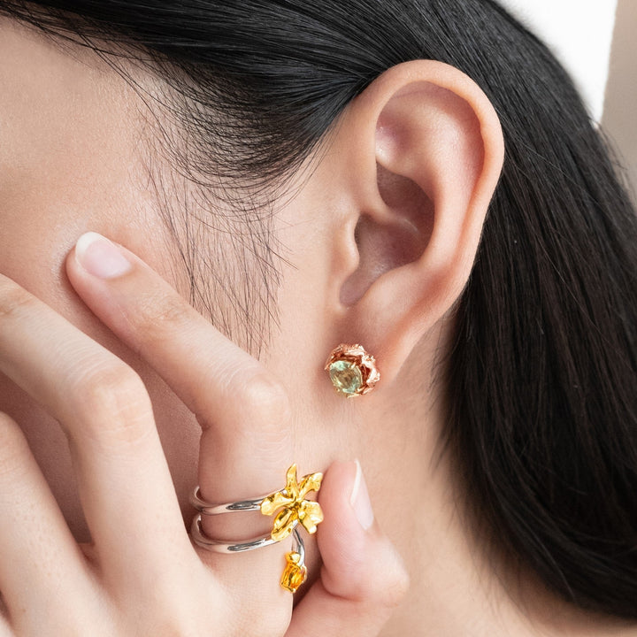 Risis Midori Earrings with Prasiolite (RG) - A person is wearing a rose gold earring with a green gemstone and is adorned with a matching ring featuring gold details and complementary stones on adjacent fingers.