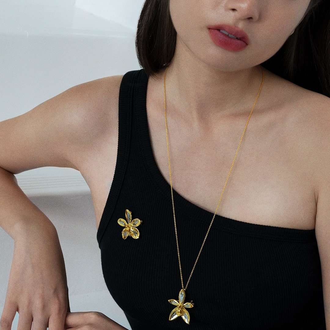 Risis Aeridovanda Vieng Ping Orchid Brooch/Pendant (PG) - A gold necklace with a flower pendant and a matching flower brooch adorn a woman in a black tank top, showcasing elegance and simplicity.