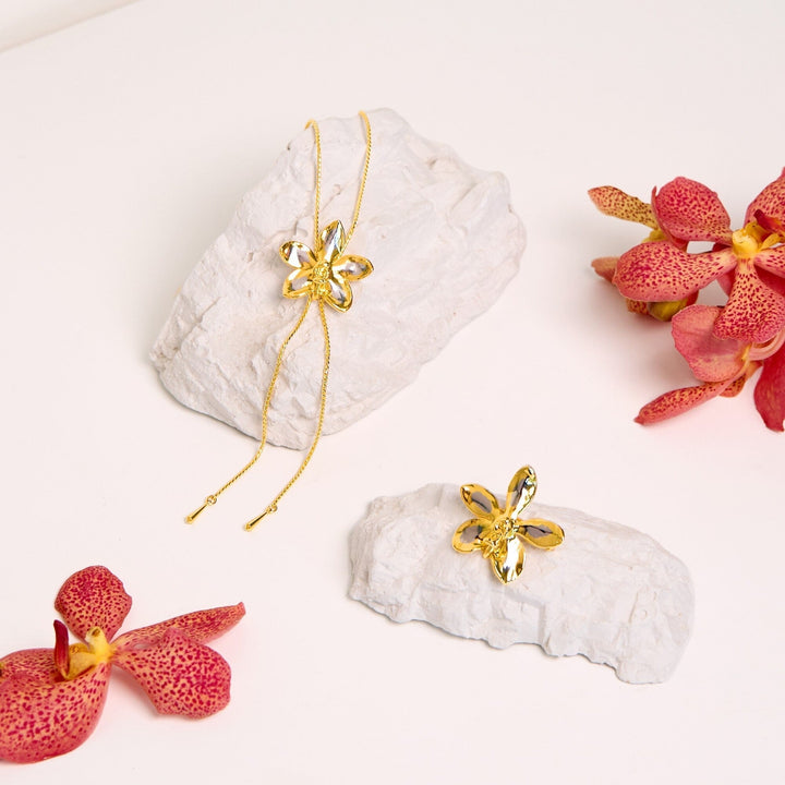 Risis Aeridovanda Vieng Ping Orchid Brooch/Pendant (PG) - A golden necklace with a flower-shaped pendant lies atop a white stone, near a matching flower-shaped earring. Pink spotted orchids complete the composition, accentuating the jewelry's elegance.