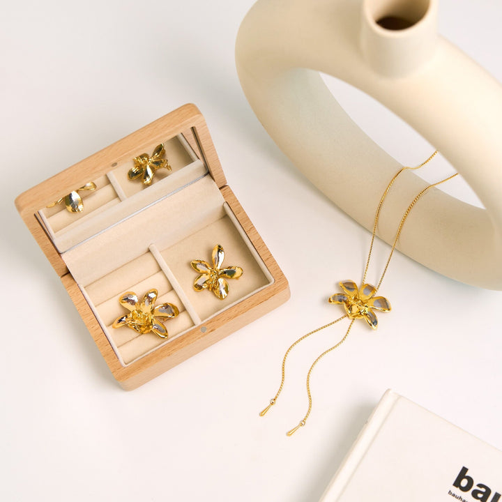 Risis Aeridovanda Vieng Ping Orchid Brooch/Pendant (PG) - A wooden jewelry box contains a set of gold-tone flower-shaped pieces: earrings and a pendant, all adorned with clear central stones on a neutral backdrop.