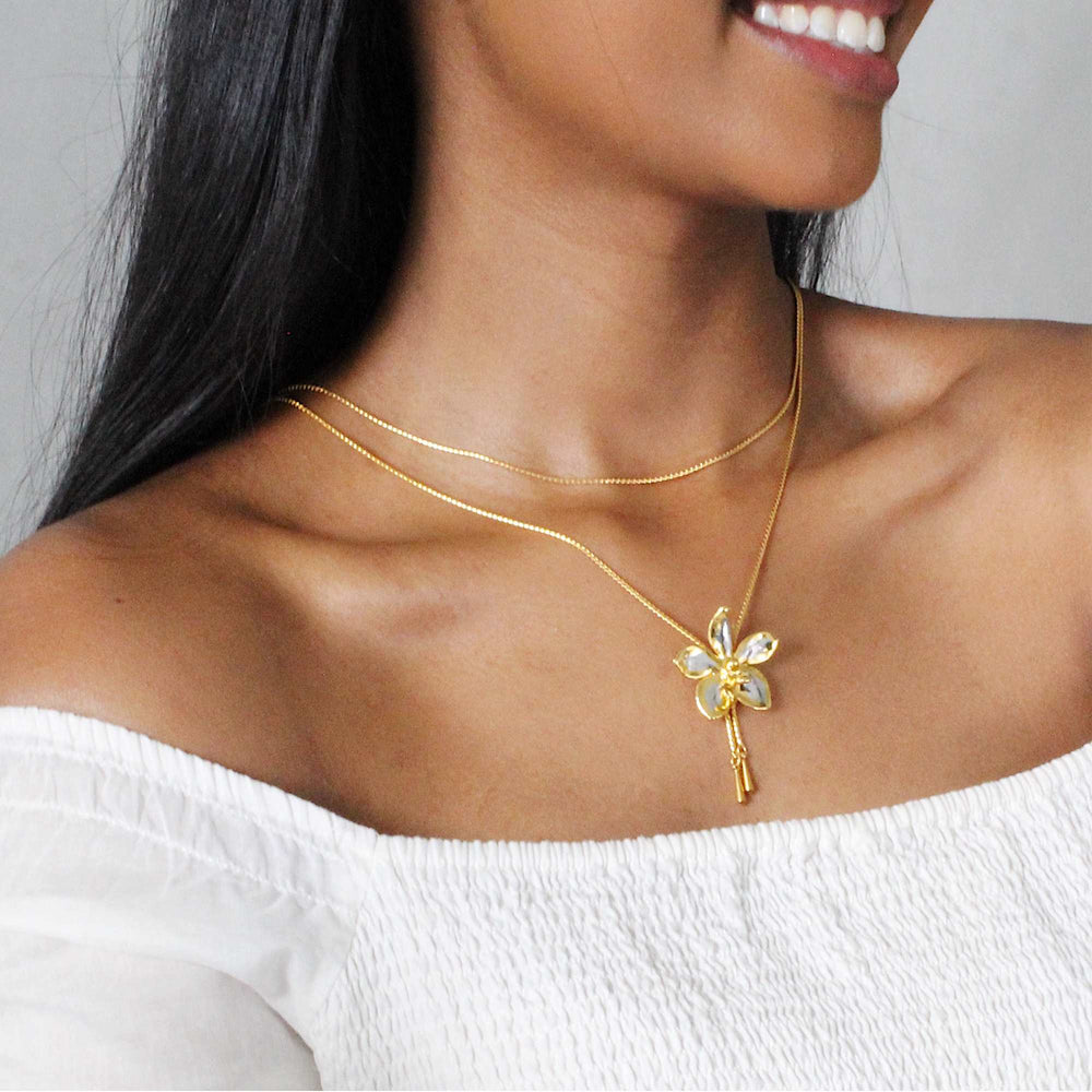 Risis Aeridovanda Vieng Ping Orchid Slider Necklace (PG) - A golden necklace with a delicate flower pendant, featuring opalescent petals and a sparkling center, adorns a woman's neck.