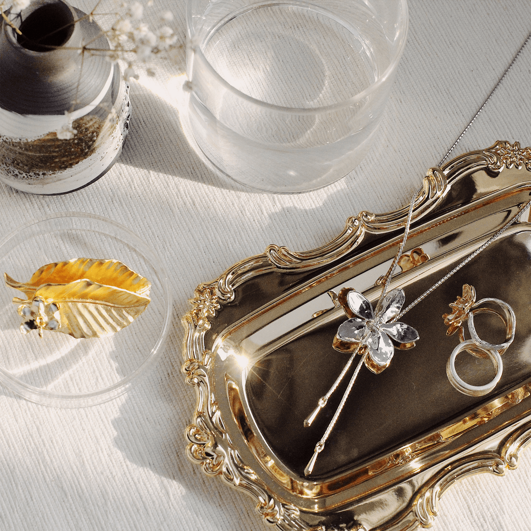 Risis Aeridovanda Vieng Ping Orchid Slider Necklace (P) - A golden tray presents a crystal flower pendant, a golden leaf brooch, and elegant rings, all gleaming under natural light.