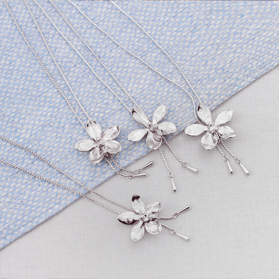 Risis Aeridovanda Vieng Ping Orchid Slider Necklace (P) - Four silver necklaces with floral pendants, featuring a design resembling five-petaled flowers, each with a textured finish, displayed on a textured surface.