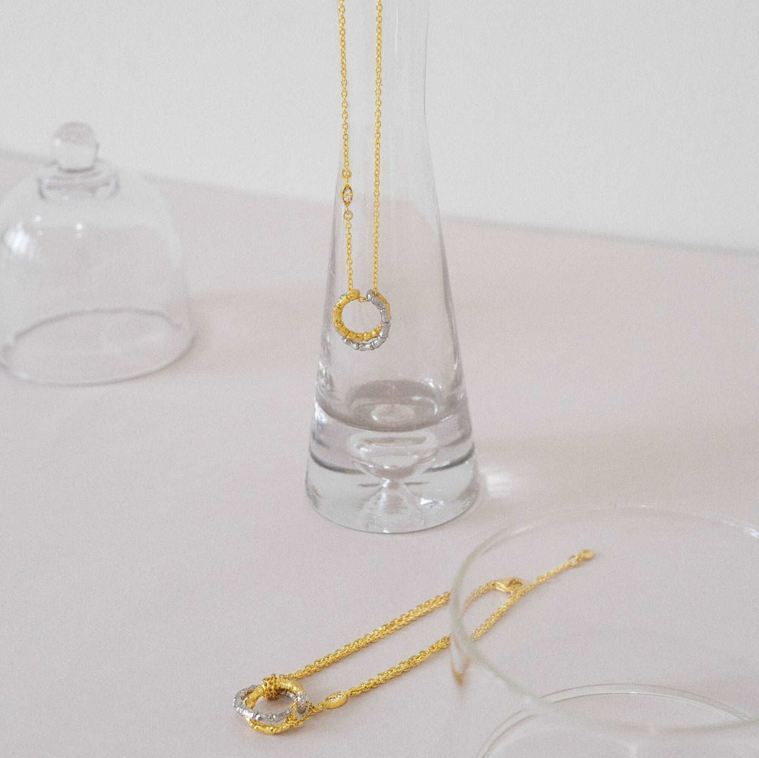 Risis Bamboo Twist Necklace with White Sapphires - A golden necklace with a circular pendant set with small gems dangles inside a clear glass vase, alongside a matching bracelet resting outside on a surface.