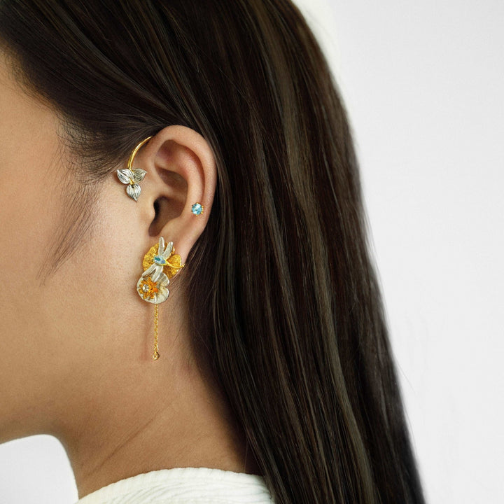 Risis Botanique Water Lily Ear Cuff - A person wearing elaborate gold and gemstone earrings, featuring floral and bee motifs, adorns an ear with a straight, sleek, brunette ponytail.