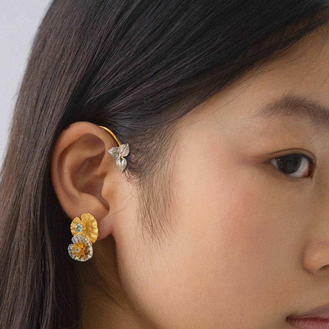 Risis Botanique Water Lily Ear Cuff - A golden floral earring with gemstone accents adorns a woman's ear, displaying intricate details and colorful craftsmanship against her dark hair.