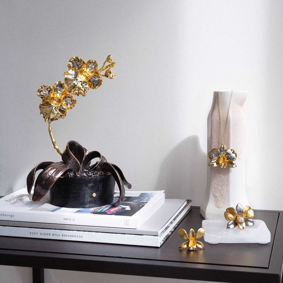 Risis Dendrobium Eleanor Chan Orchid Necklace (PG) - A gold-colored metal sculpture of delicate flowers rests on books, accompanied by similar design elements in jewelry, conveying a theme of floral artistry and elegance.
