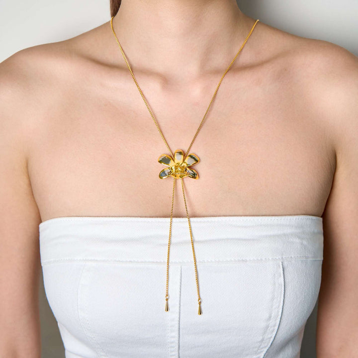 Risis Dendrobium Thong Chai Orchid Slider Necklace (PG) - A golden necklace with a delicate bow pendant adorned with translucent green gemstones hangs centered on a person's upper chest area.