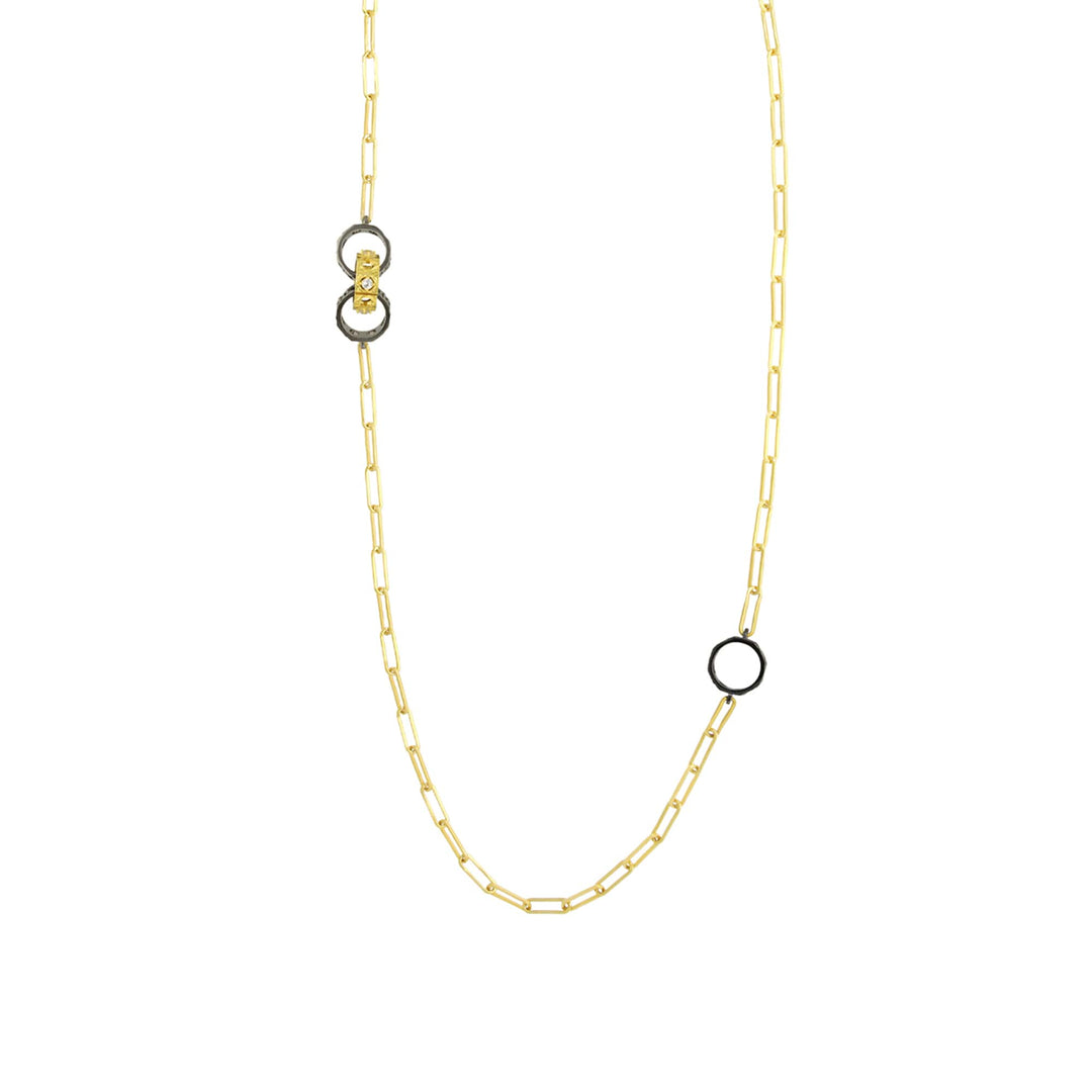 Risis Entwined Rattan Necklace - A gold necklace with a delicate chain and a transparent gemstone pendant adorns the neck of a person wearing a pastel top. Matching golden earrings peak out subtly.