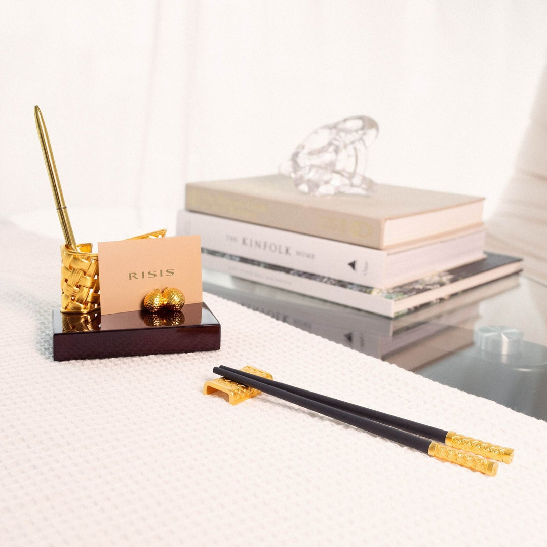 Risis Entwined Rattan Pen and Name Card Holder - A golden pen holder and a cardholder with intricate designs rest on a dark wooden base, accompanied by elegant black chopsticks adorned with golden elements on their ends.