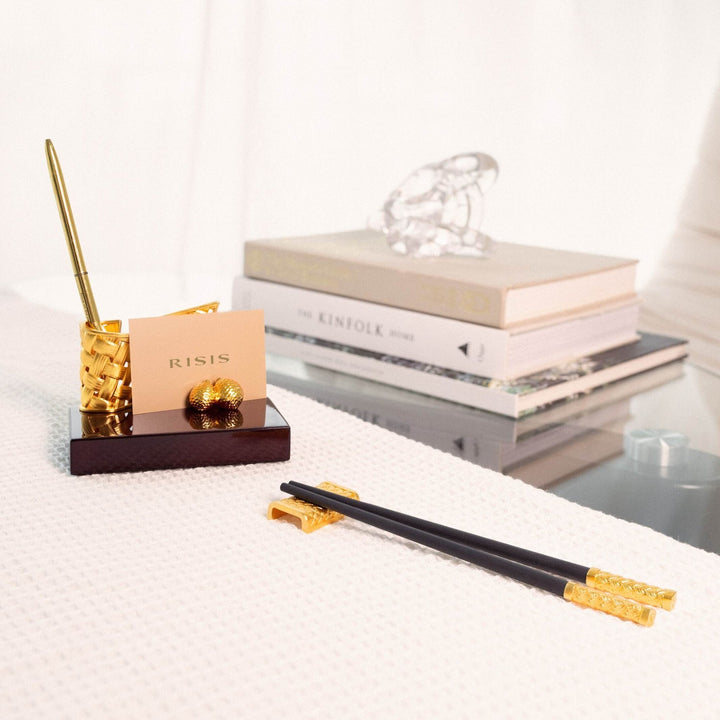 Risis Entwined Rattan Pen and Name Card Holder - A golden pen holder and a cardholder with intricate designs rest on a dark wooden base, accompanied by elegant black chopsticks adorned with golden elements on their ends.
