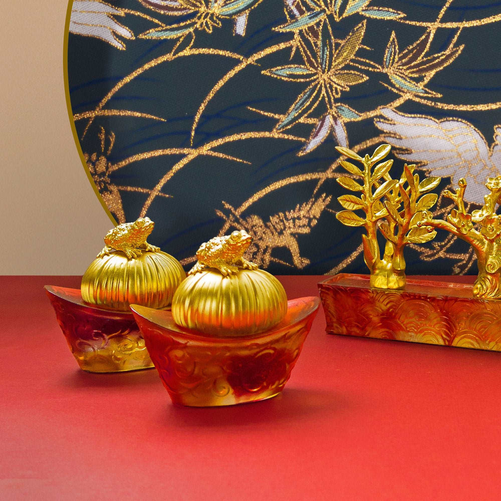 Risis Fortune Toad on Red Yuan Bao - Two ornate, gold-colored objects resembling pumpkin-shaped boxes with frog figurines on top are set on amber-hued, translucent bases with swirling patterns, emanating luxury and artistic flair.