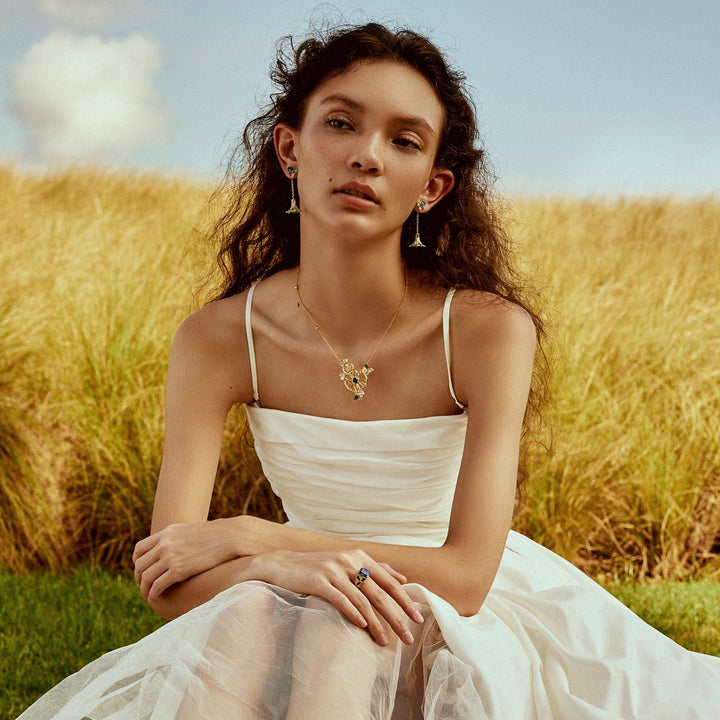 Risis Garden Odyssey Drop Earrings - A woman adorned with elegant jewelry; gold necklace with intricate charm, dangle earrings with green gemstones, and a ring with a large blue gemstone on her finger.
