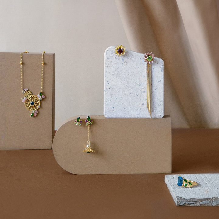 Risis Garden Odyssey Drop Earrings - Elegant jewelry—pendants and earrings—with intricate designs featuring gold elements and colorful gemstones, artistically arranged on beige stands.
