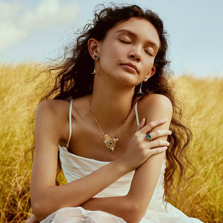 Risis Garden Odyssey Drop Earrings - A woman adorned with elegant jewelry, wearing a white dress, is featured in an outdoor setting. The jewelry comprises a delicate necklace with a floral pendant and green accents, matching drop earrings, and a statement ring with a blue gemstone.