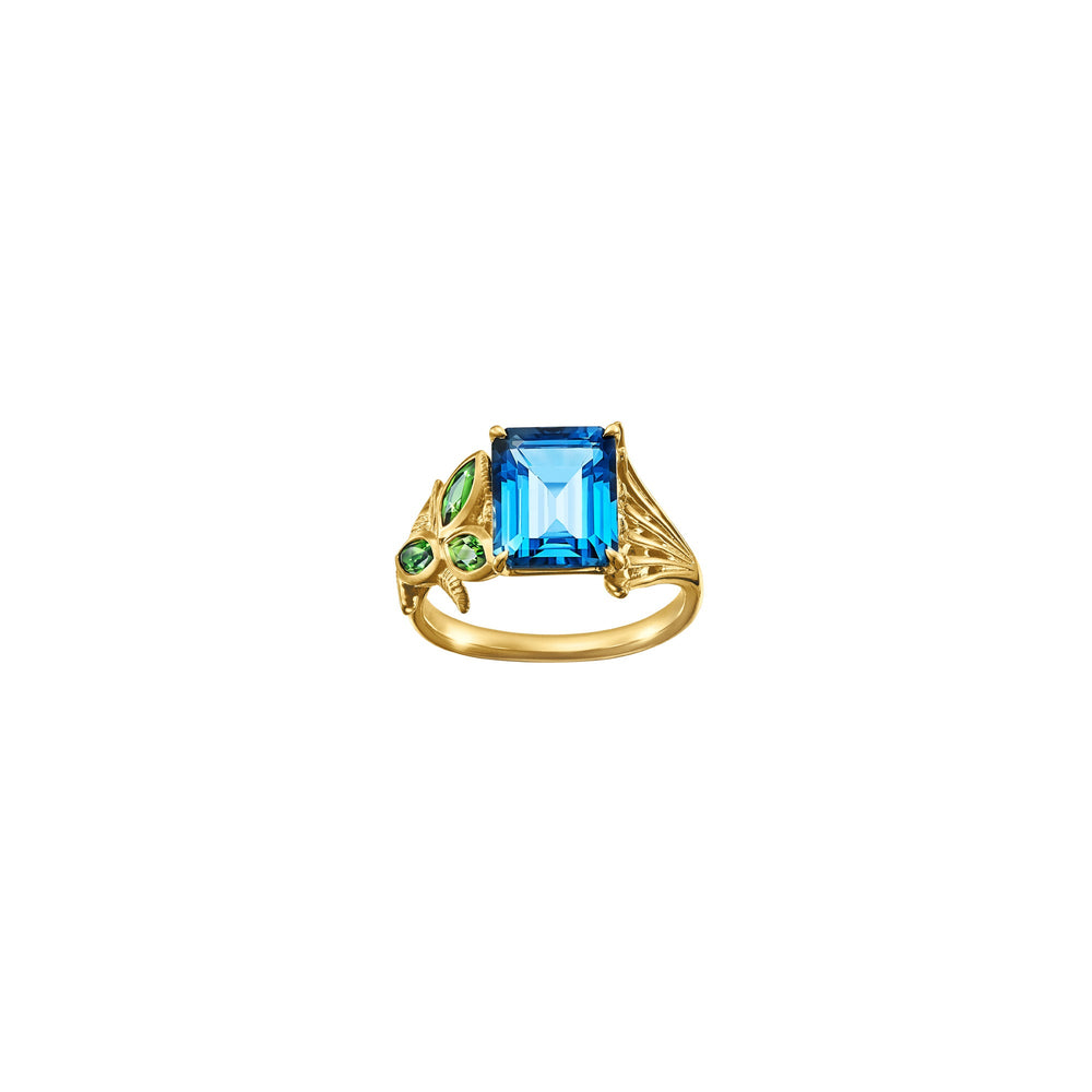 Risis Garden Odyssey Medley Ring - A golden ring features a prominent rectangular blue gemstone at the center, complemented by smaller green gemstones arranged in a leaf-like pattern on one side.
