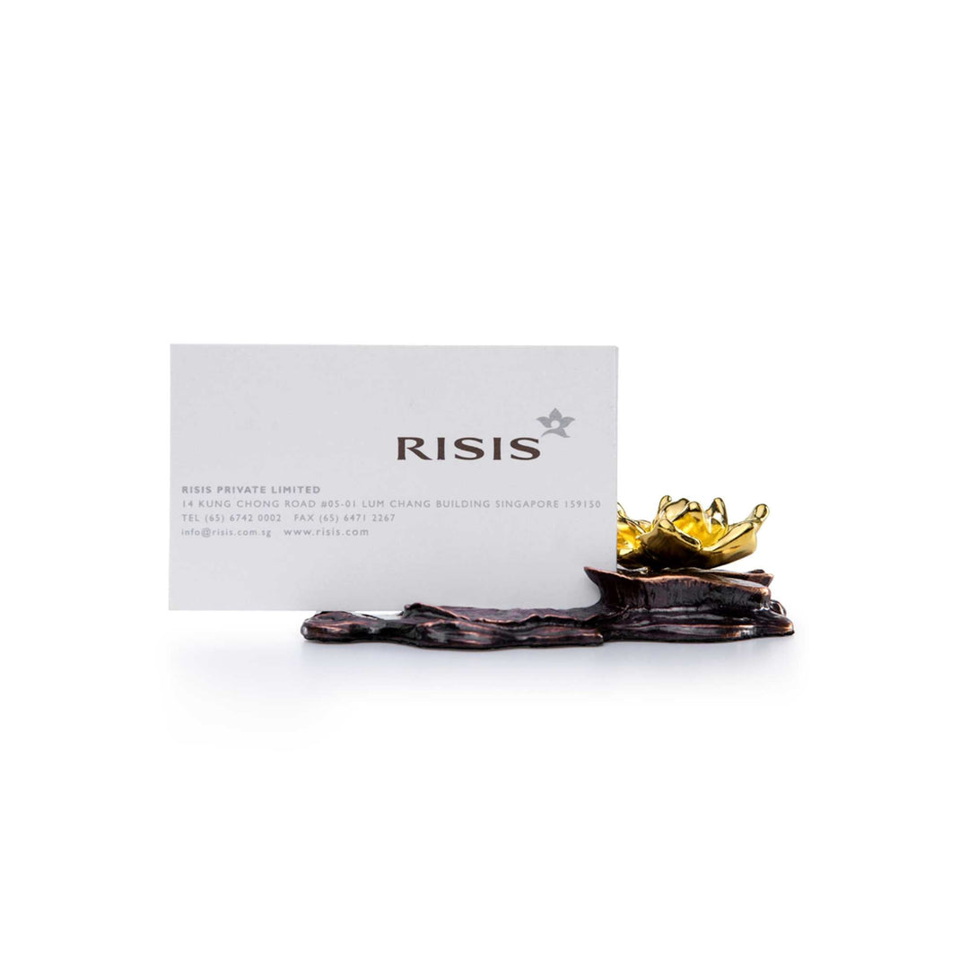 Risis Lotus Name Card Holder - A gold-tinted lotus flower sculpture resting on a textured, dark platform, accompanied by a white business card with text and a logo.