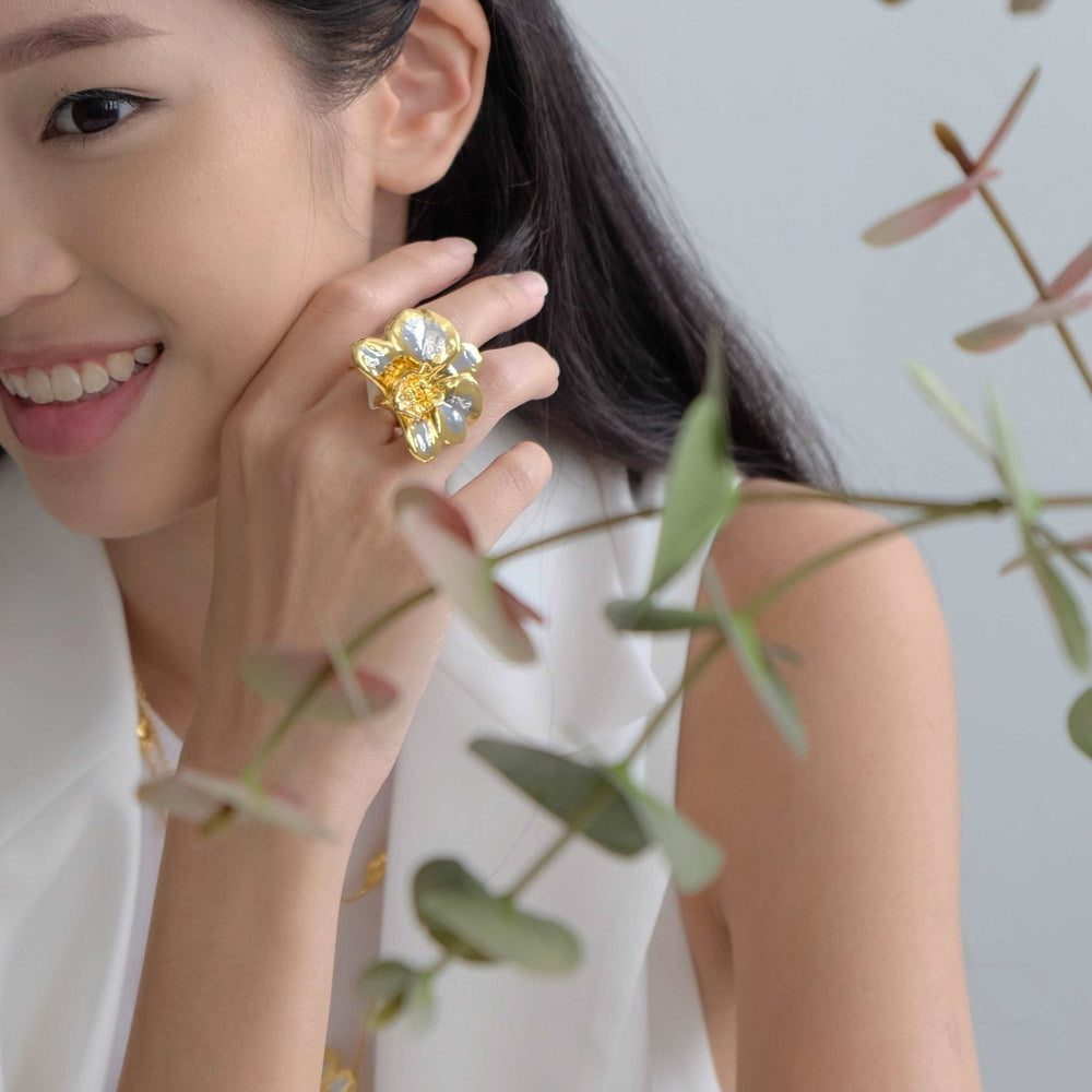 Risis Merry Blossom Ring (PG) - A person is displaying a large, gleaming gold ring with a floral design, adorned with translucent and yellow petals on their finger.