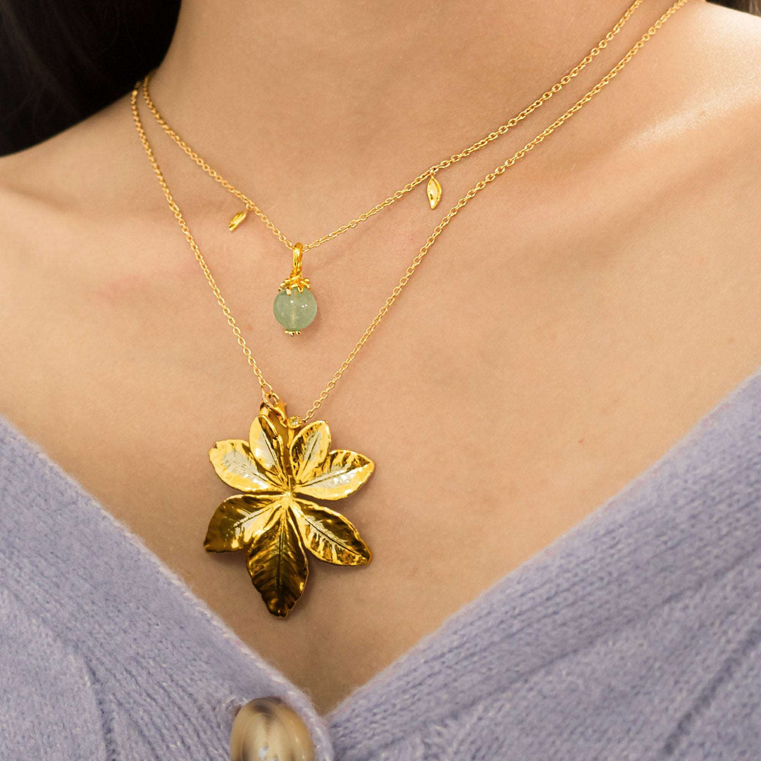 Risis Morning Glory Leaf Necklace with Aquamarine and Topaz - A golden necklace featuring a large textured leaf pendant and a smaller translucent green bead, layered above a delicate chain with tiny leaf charms.