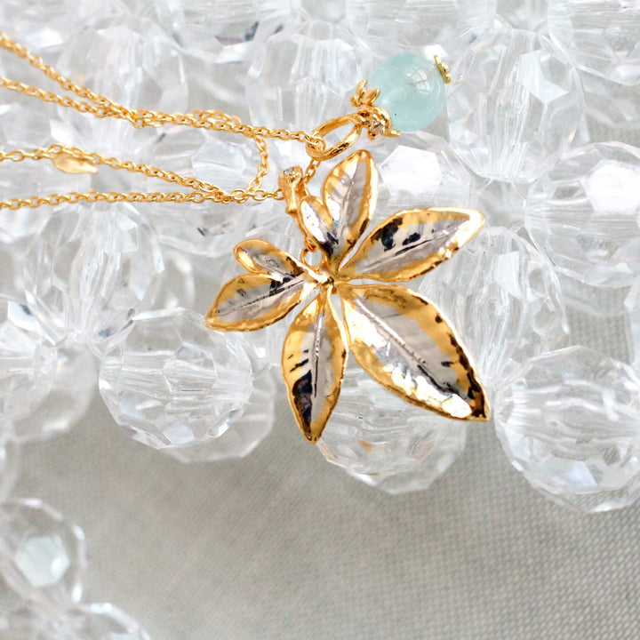 Risis Morning Glory Leaf Necklace with Aquamarine and Topaz - A gold necklace with a detailed, petal-shaped pendant and a small, pale green bead dangles elegantly above a sparkling surface.