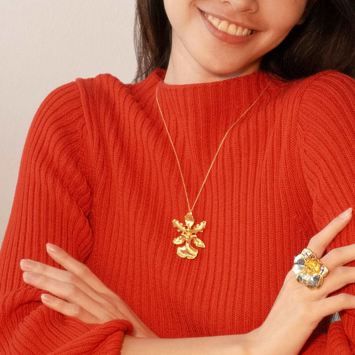 Risis Oncidium Haematochilum Orchid Necklace - A smiling person wears a red sweater, a gold necklace with a floral pendant, and a large multicolored gemstone ring.