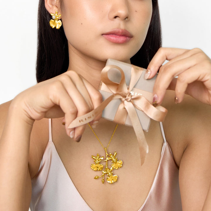 Risis Oncidium Shower Earrings (G) - A person showcases gold jewelry: floral earrings and a matching necklace, complemented by a muted gold gift box with a satin ribbon in their hands.