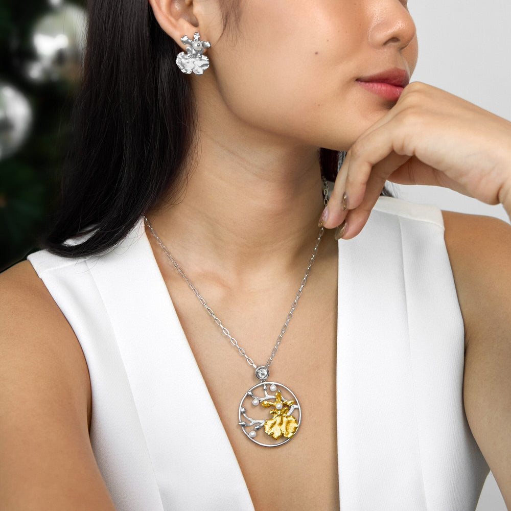 Risis Oncidium Shower Necklace - A woman showcases a silver necklace with a circular pendant featuring a golden element and small gemstones, paired with matching silver floral earrings.