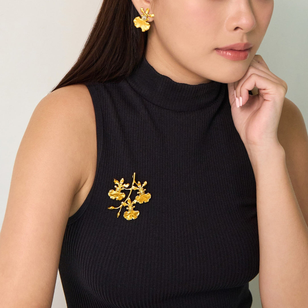 Risis Oncidium Shower Spray Brooch/Pendant (G) - A golden floral earring and brooch set adorn a person's ear and black turtleneck. The jewelry features detailed petals and a realistic, textured design.