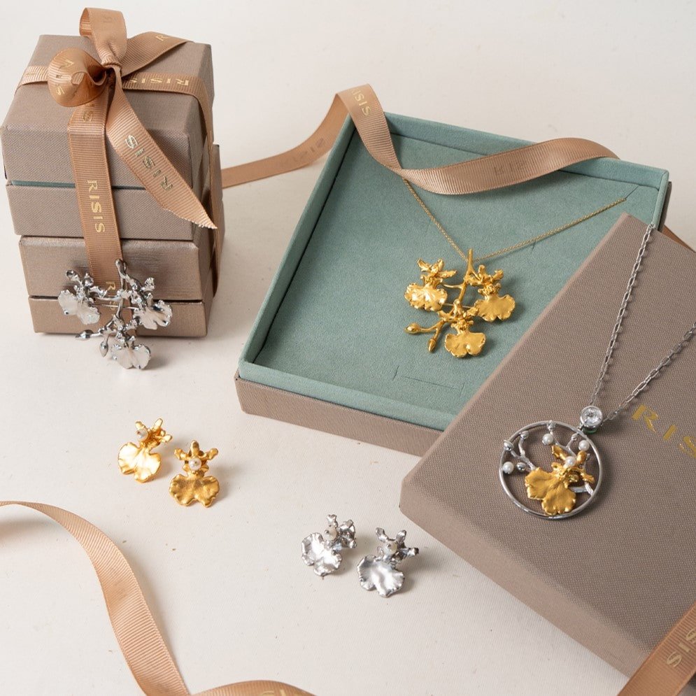 Risis Oncidium Shower Spray Brooch/Pendant (G) - Assorted jewelry pieces including a silver snowflake pendant, gold floral earrings, and silver blossom earrings are displayed alongside elegant gift boxes with beige ribbons.