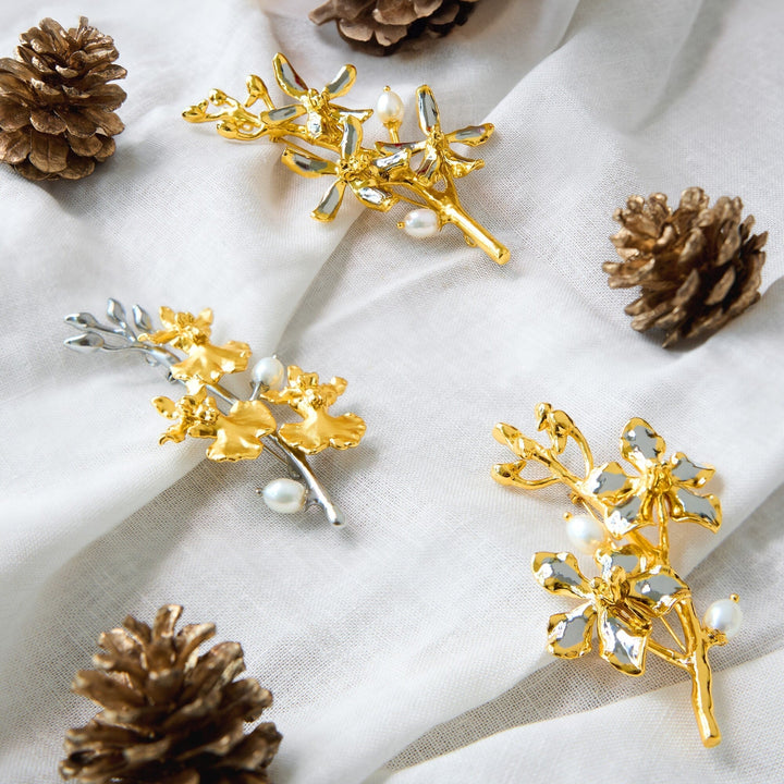 Risis Oncidium Symphony Brooch with Pearls - Golden and silver-colored floral brooches with pearl embellishments rest on a textured white fabric, accompanied by pinecones, presenting an elegant and natural motif.