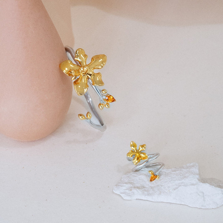 Risis Orchid Spiral Bangle - A hand displays a silver ring with golden flower embellishments and small red stones, complementing a pair of matching floral earrings.
