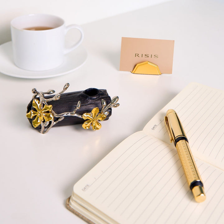 Risis Orchid Spiral Charcoal Pen and Name Card Holder - A sculptural black jewelry piece adorned with silver and gold floral accents sits beside a gold-toned pen on a lined notebook.