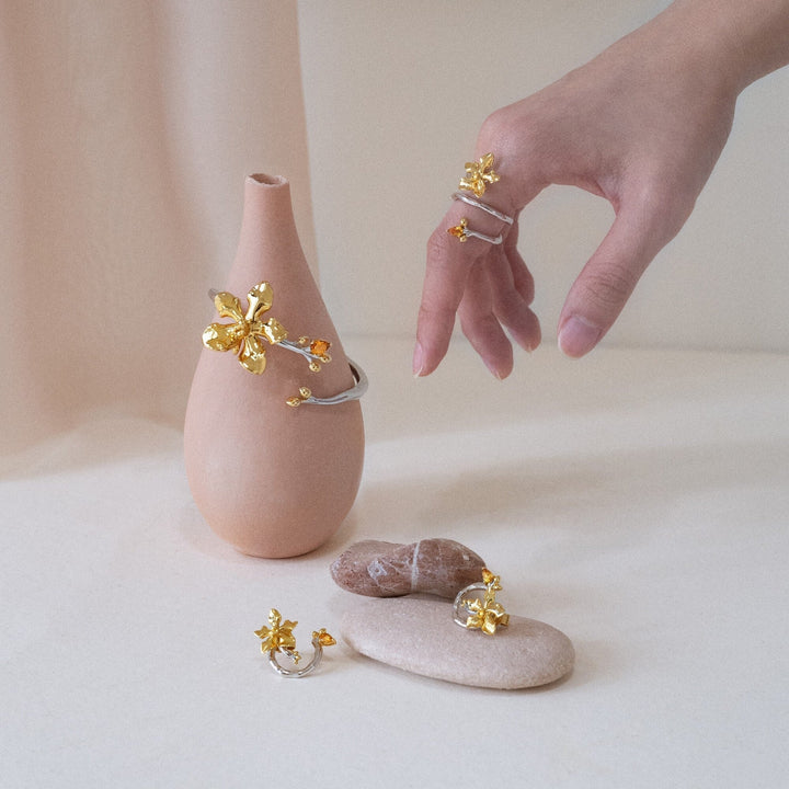 Risis Orchid Spiral Earrings - A woman showcases gold jewelry, including floral-design earrings, a bracelet, and a ring, with an elegant and modern aesthetic.