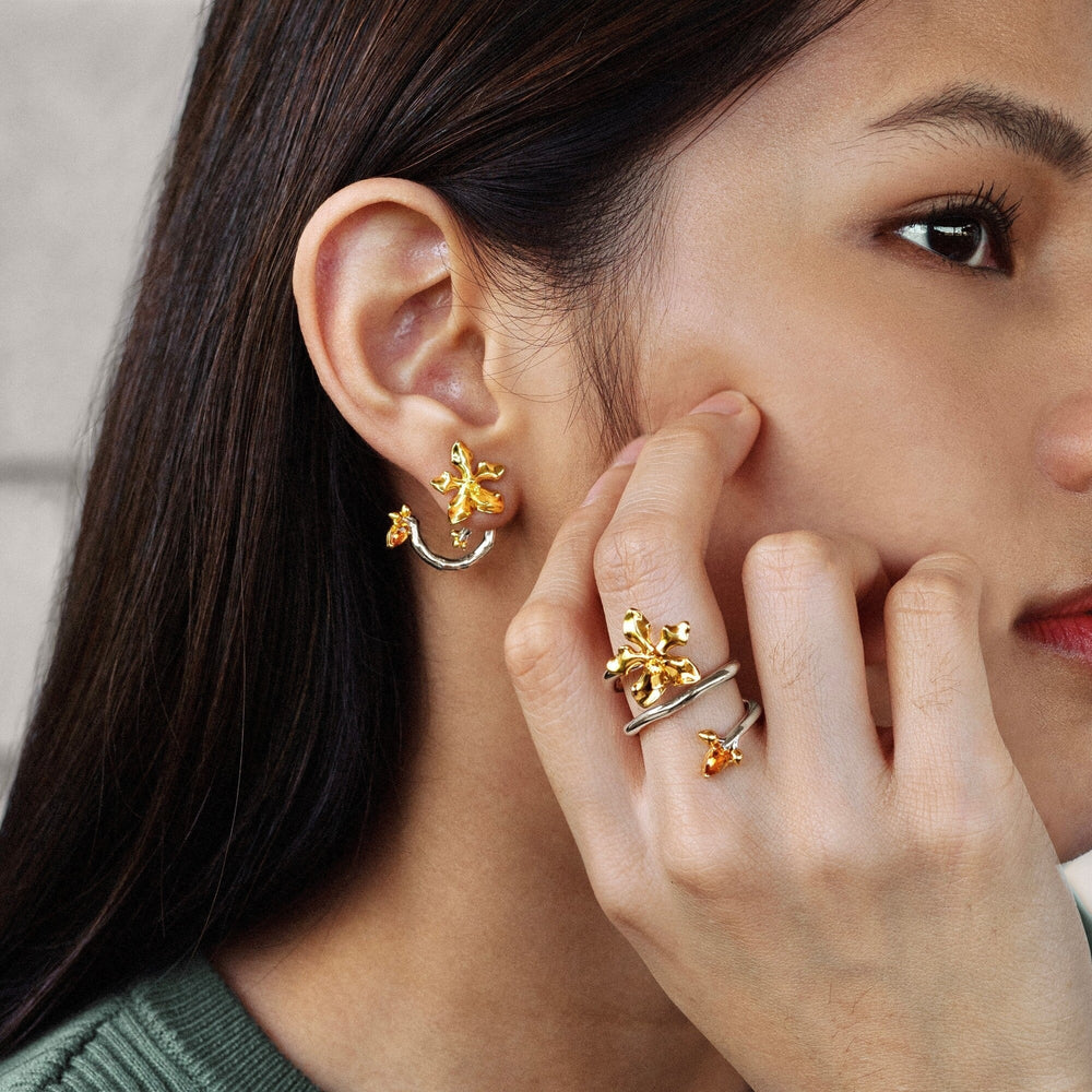 Risis Orchid Spiral Earrings - A woman is wearing intricate gold and silver floral jewelry, including a cuff earring and rings, highlighting elegance against her skin and dark hair.