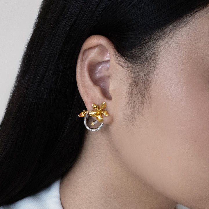 Risis Orchid Spiral Earrings - Gold and silver jewelry with floral motifs gracefully rest on smooth, beige stones. The pieces exude elegance and craftsmanship through their polished finish and intricate designs.